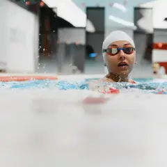 Kobieta w czepku i okularach pływackich w basenie, z wodą rozpryskującą się wokół.