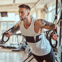 Mężczyzna z tatuażami ćwiczy na taśmach TRX. Zastanawiasz się, ile trwa trening na siłowni? Ten intensywny wysiłek to klucz do sukcesu.