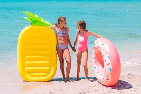 Dwie dziewczynki w strojach kąpielowych stoją na plaży obok dmuchanej ananasa i pączka.