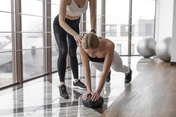 Trening w domu: kobieta w pozycji deski z piłką, druga osoba pomaga. Efektywny trening nie musi trwać długo.