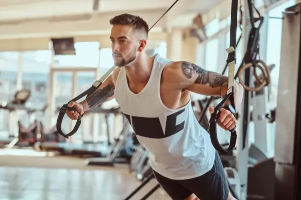 Mężczyzna z tatuażami ćwiczy na taśmach TRX. Zastanawiasz się, ile trwa trening na siłowni? Ten intensywny wysiłek to klucz do sukcesu.